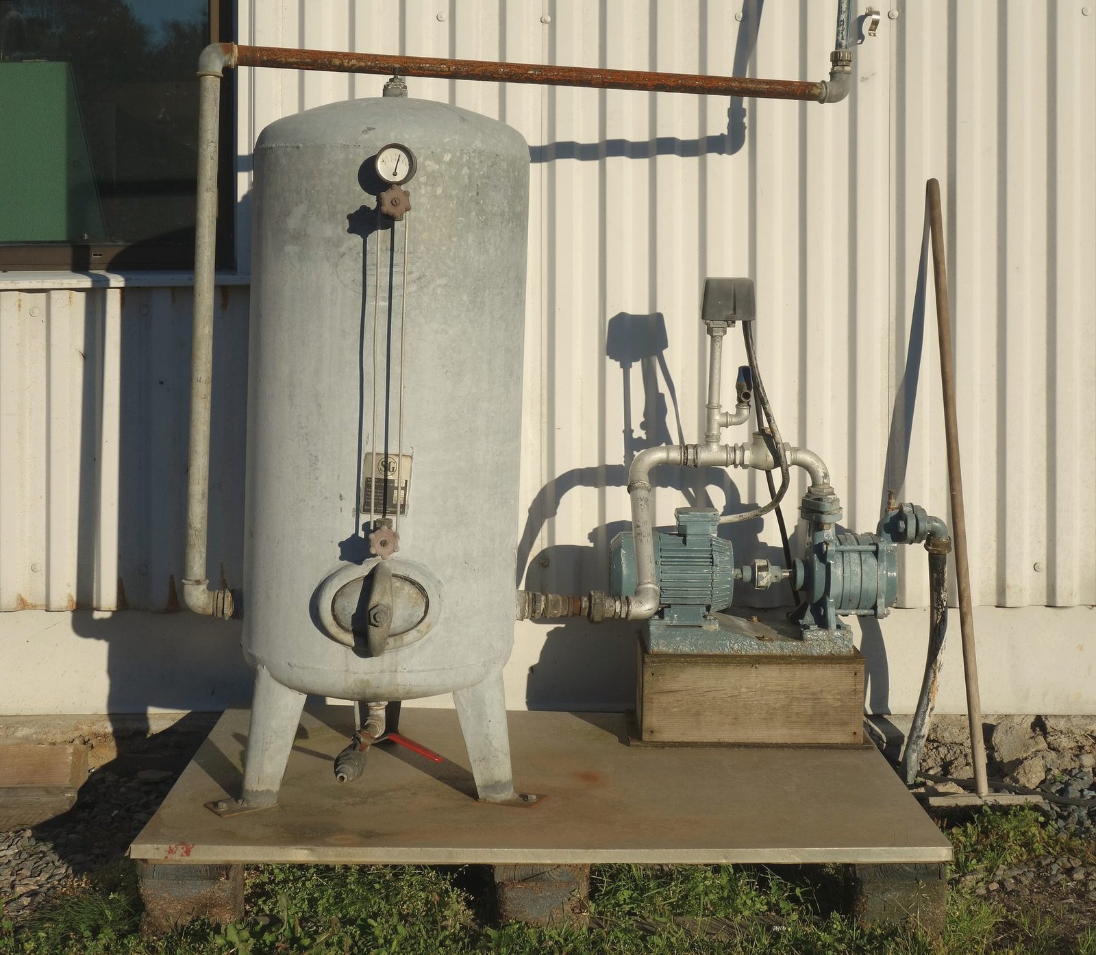 A grey pressure tank and an electric pump on a concrete pad beside a metal building